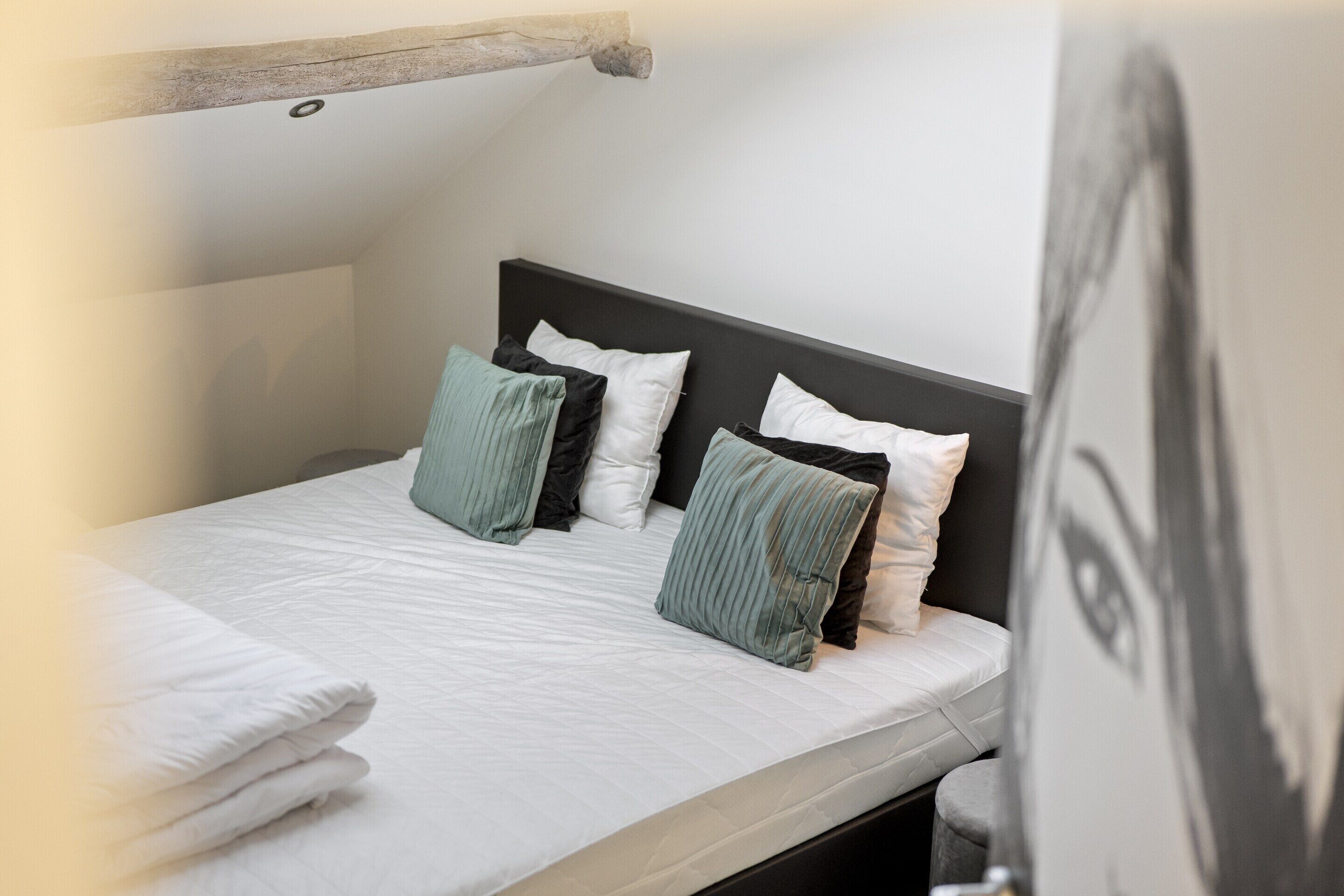 Una cama bien hecha con ropa blanca y varias almohadas en una habitación con techo inclinado y cabecera oscura.