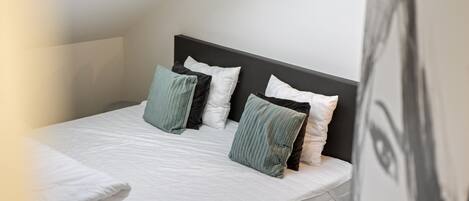 Una cama bien hecha con ropa blanca y varias almohadas en una habitación con techo inclinado y cabecera oscura.