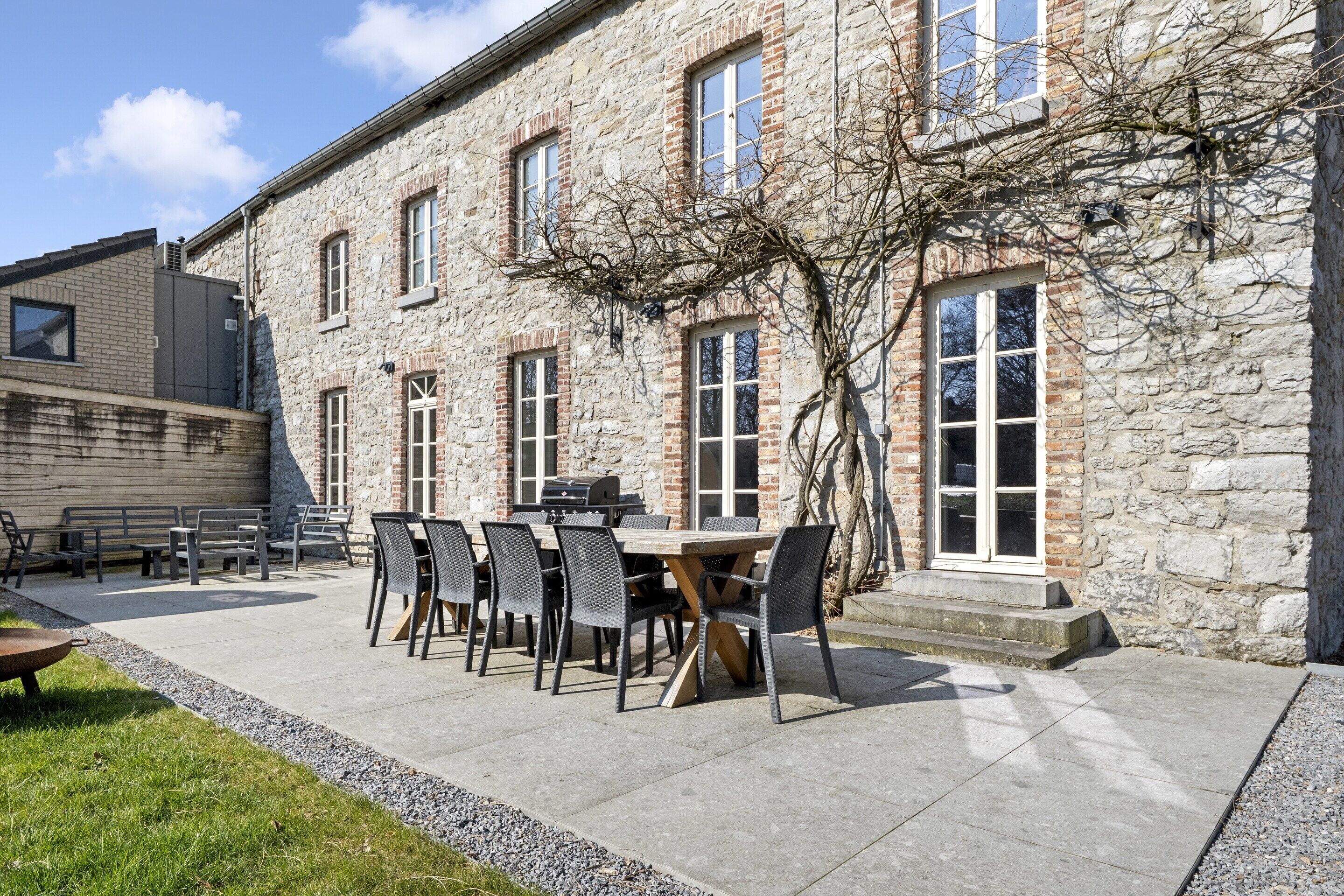 A stone building with a patio area featuring a table and chairs, a grill, and a small garden.
