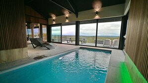 Una piscina interior con vistas al exterior, un área de descanso con sillones y un porche cubierto.