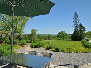 Um pátio externo com mesa e cadeiras, um guarda-sol verde e vista para um campo gramado e árvores.