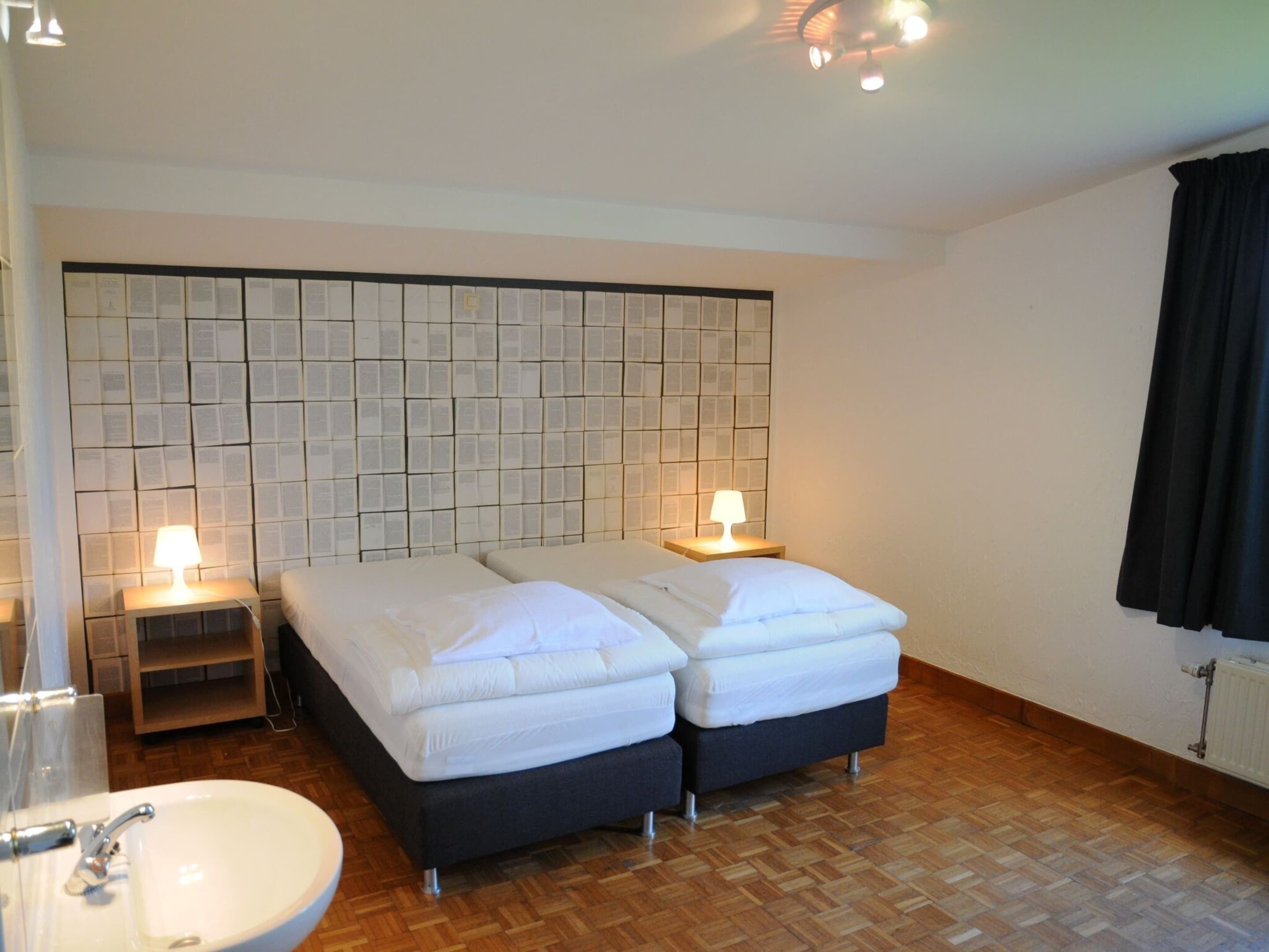 A hotel room with two beds, a bedside table with a lamp, a wooden floor, a white sink, and a tiled wall with a pattern.