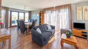 A modern living room with a grey sofa, wooden coffee table, and a TV on a wooden stand. There is a dining area with a table and chairs, and a bookshelf filled with books. - Holiday Home in Borlon With Terrace (Durbuy)