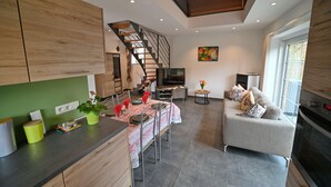 A modern kitchen with wooden cabinets, a dining area with a table set for four, a staircase, a sofa, and a TV in a living room.