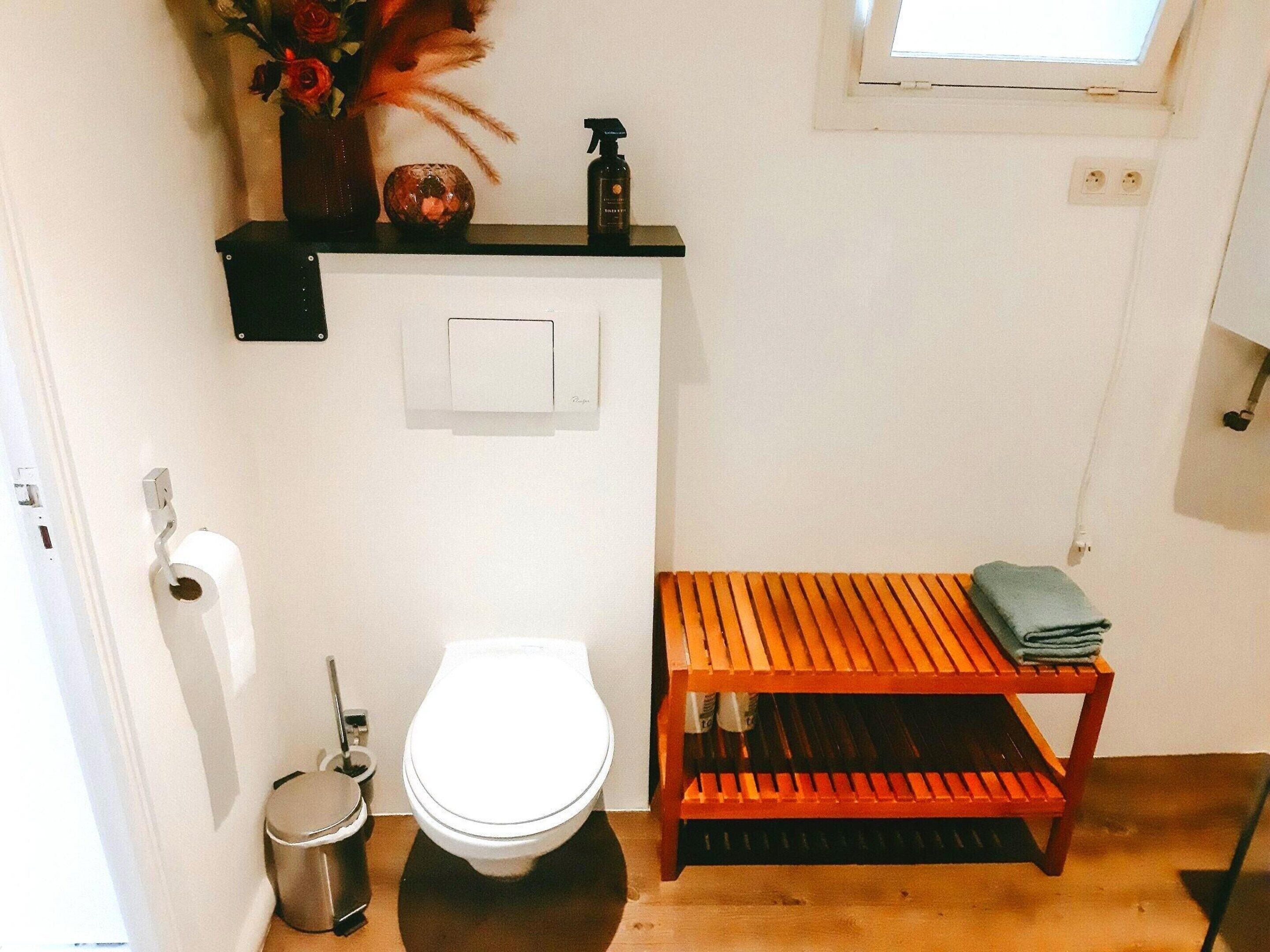 A small bathroom with a toilet, a sink, a shelf with toiletries, and a window.