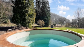 Une piscine ronde en bois, dont l’intérieur est d’un bleu clair, entourée d’une terrasse en bois et d’une zone herbeuse agrémentée d’arbres, avec une montagne en arrière-plan.