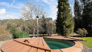 Un bain à remous en bois, doté d’un bord incurvé, entouré d’une terrasse carrelée, installé dans un jardin aménagé avec des arbres et sous un ciel dégagé.