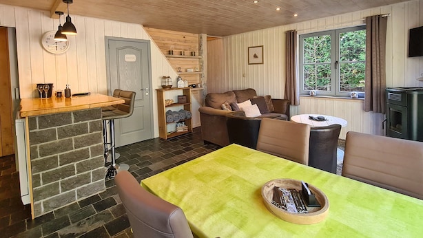 En mysig stuga med träpanel i taket, ett matbord med en grön duk, en soffa, en öppen spis och ett fönster med gardiner.