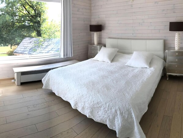 A bedroom with a large bed, two bedside tables, a bench, and a window with a view of trees.