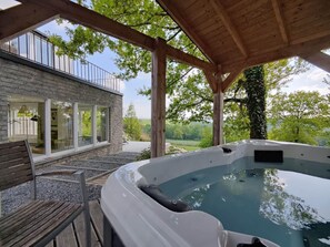 A hot tub under a wooden pergola with a view of a stone building and trees.
