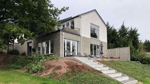 Een modern huis met twee verdiepingen, een ruim balkon, een overdekt terrasgedeelte met lounge stoelen, en een trap naar de ingang.