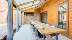 A long wooden table set for dining with chairs, a brick wall, and a glass ceiling. - Beautiful Holiday Home in Merksplas With Garden (Merksplas)