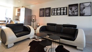 A modern living room with a black and white sofa, a coffee table, and a rug. There are framed pictures on the wall and a kitchen in the background. - Rochehaut Hideaway With Tub (Bouillon)