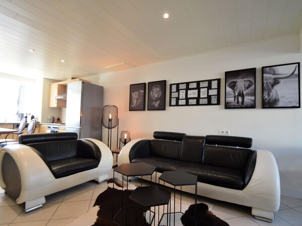 Una sala de estar moderna con un sofá blanco y negro, una mesa de centro y una alfombra. Hay cuadros enmarcados en la pared y una cocina al fondo.