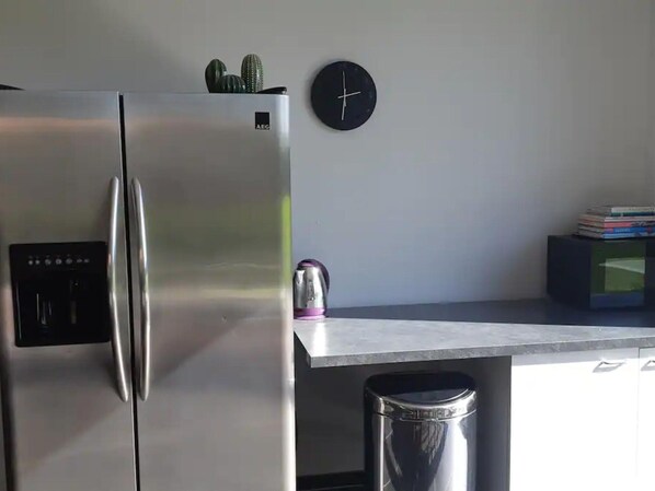 A kitchen with a stainless steel refrigerator, a kettle, a trash bin, and a clock on the wall.