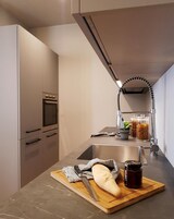 Una cucina moderna con forno integrato, lavello in acciaio inox e un tagliere di legno con pane e un vasetto di marmellata.