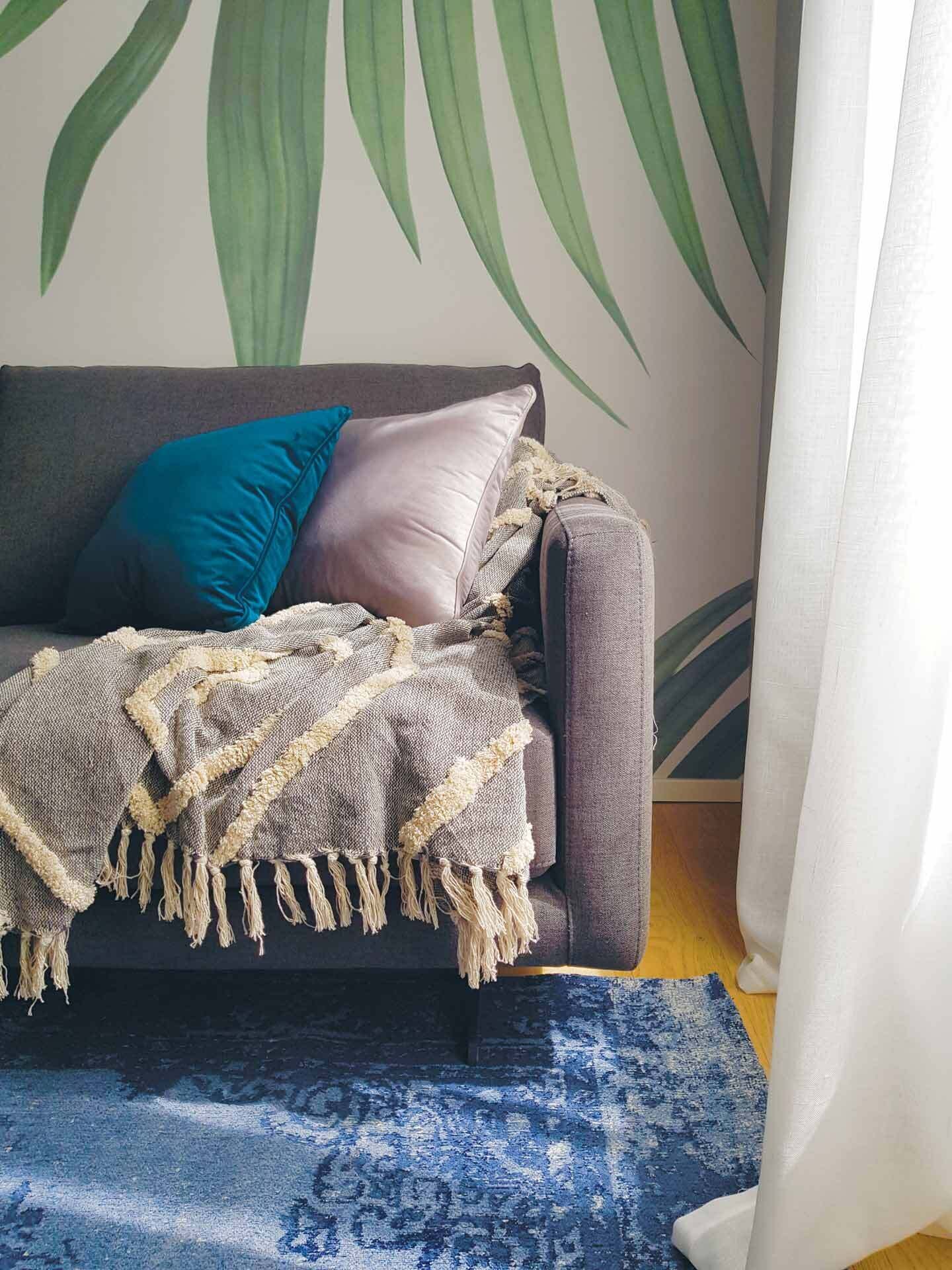 Un sofá gris con una manta beige y dos almohadones, un tapete azul estampado y un mural de una hoja verde grande.