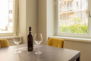 Un tavolo con due bicchieri da vino e una bottiglia, una sedia gialla e una finestra con vista su edifici e vegetazione.