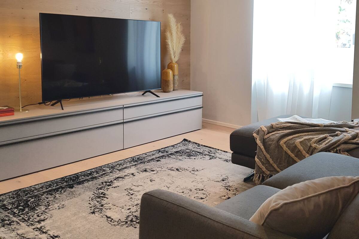 A modern living room with a flat-screen TV on a wooden wall, a grey sofa, and a patterned rug.