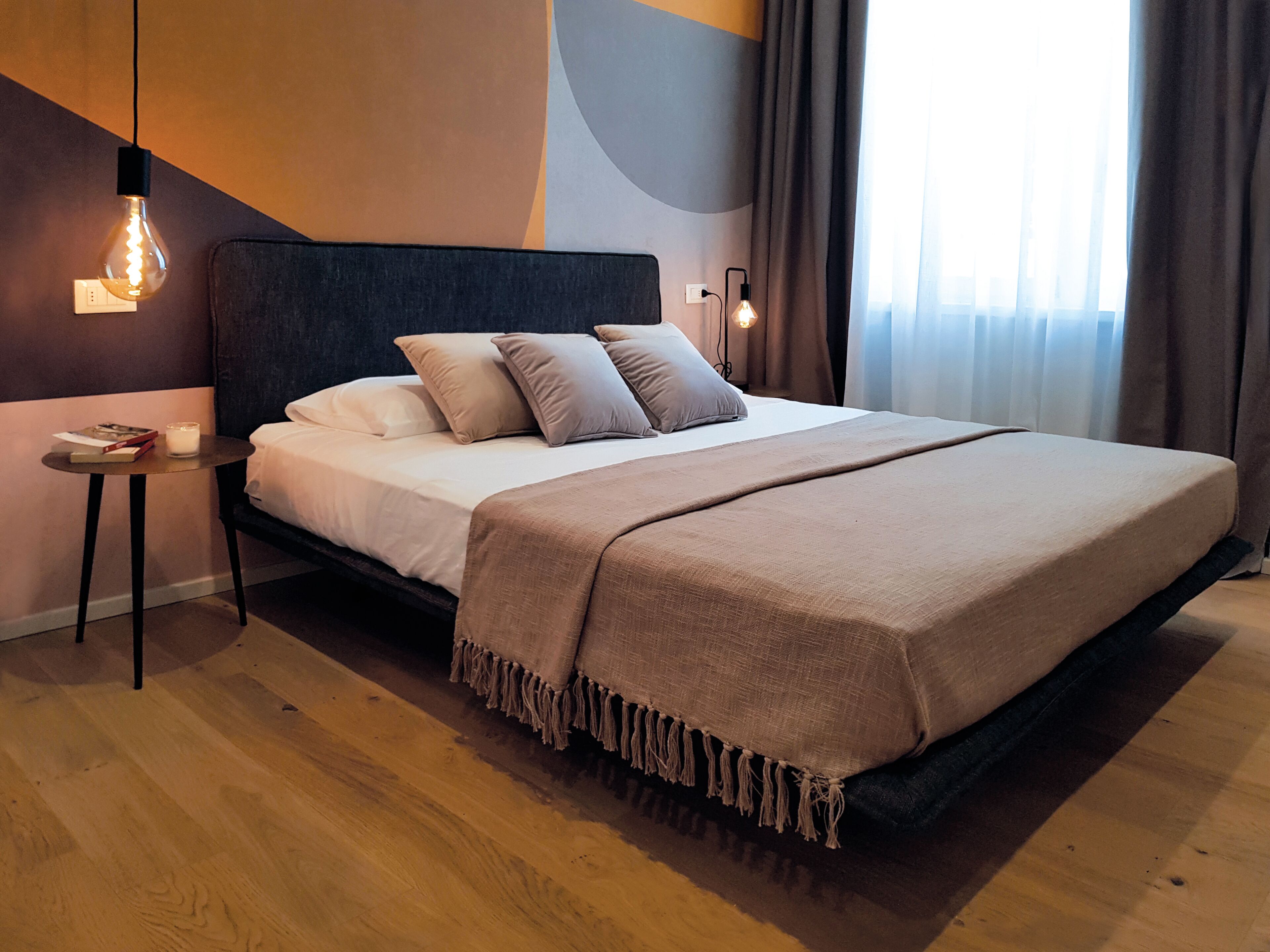 A modern hotel room with a large bed, a bedside table with a book and candle, and two wall-mounted lights.
