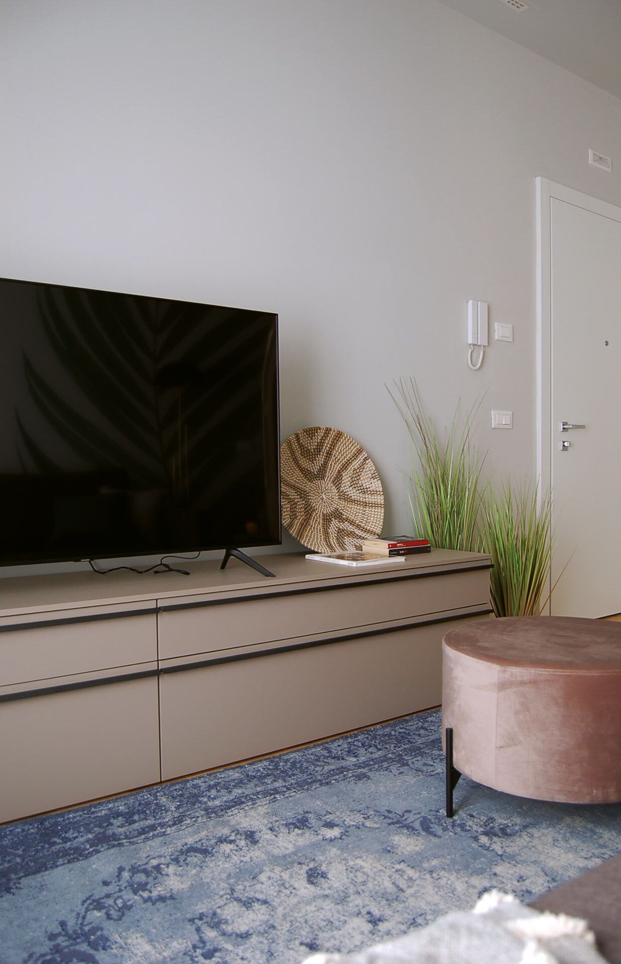 A modern living room with a flat-screen TV on a cabinet, a round woven wall decor, a potted plant, and a round ottoman on a blue patterned rug.