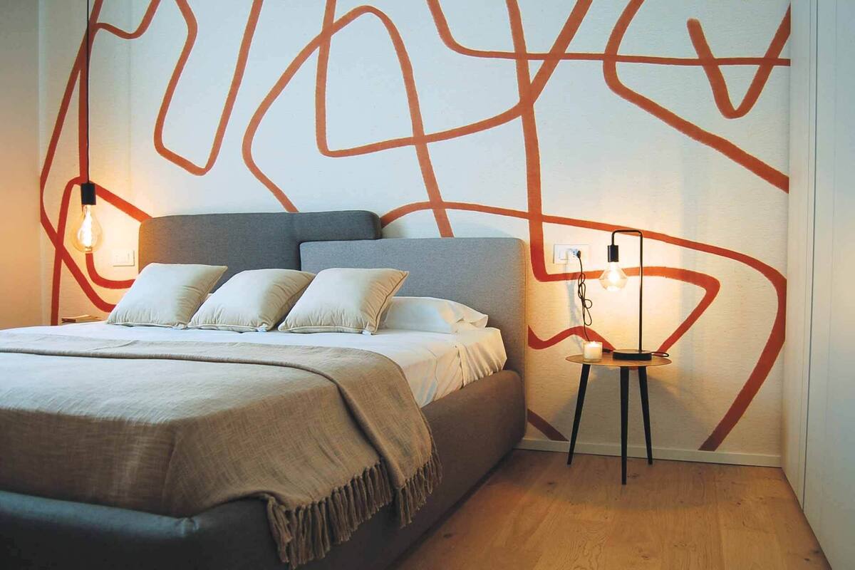 A modern bedroom with a large bed, a bedside table with a lamp, and a wall with abstract red lines.