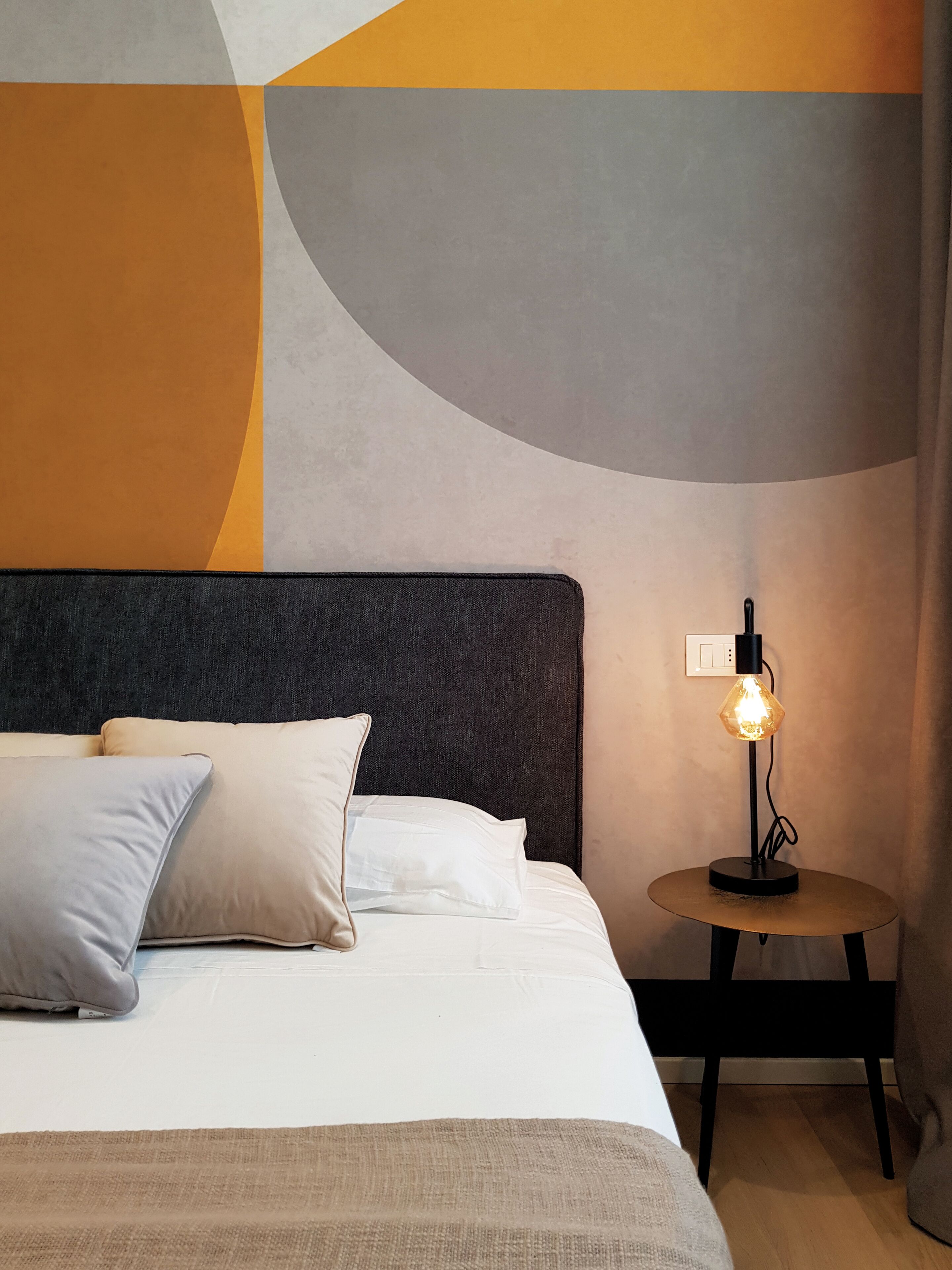 Una cama bien tendida con almohadas, una mesita redonda con una lámpara y una pared con patrones geométricos abstractos.