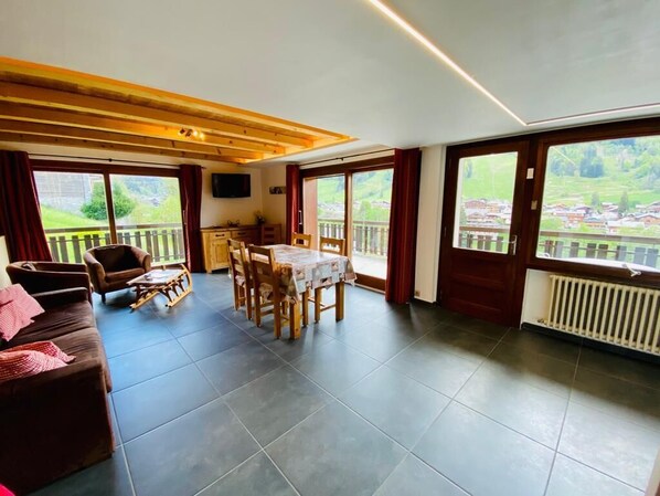 A spacious living area with a dark tiled floor, wooden furniture, and large windows offering a view of a green landscape.