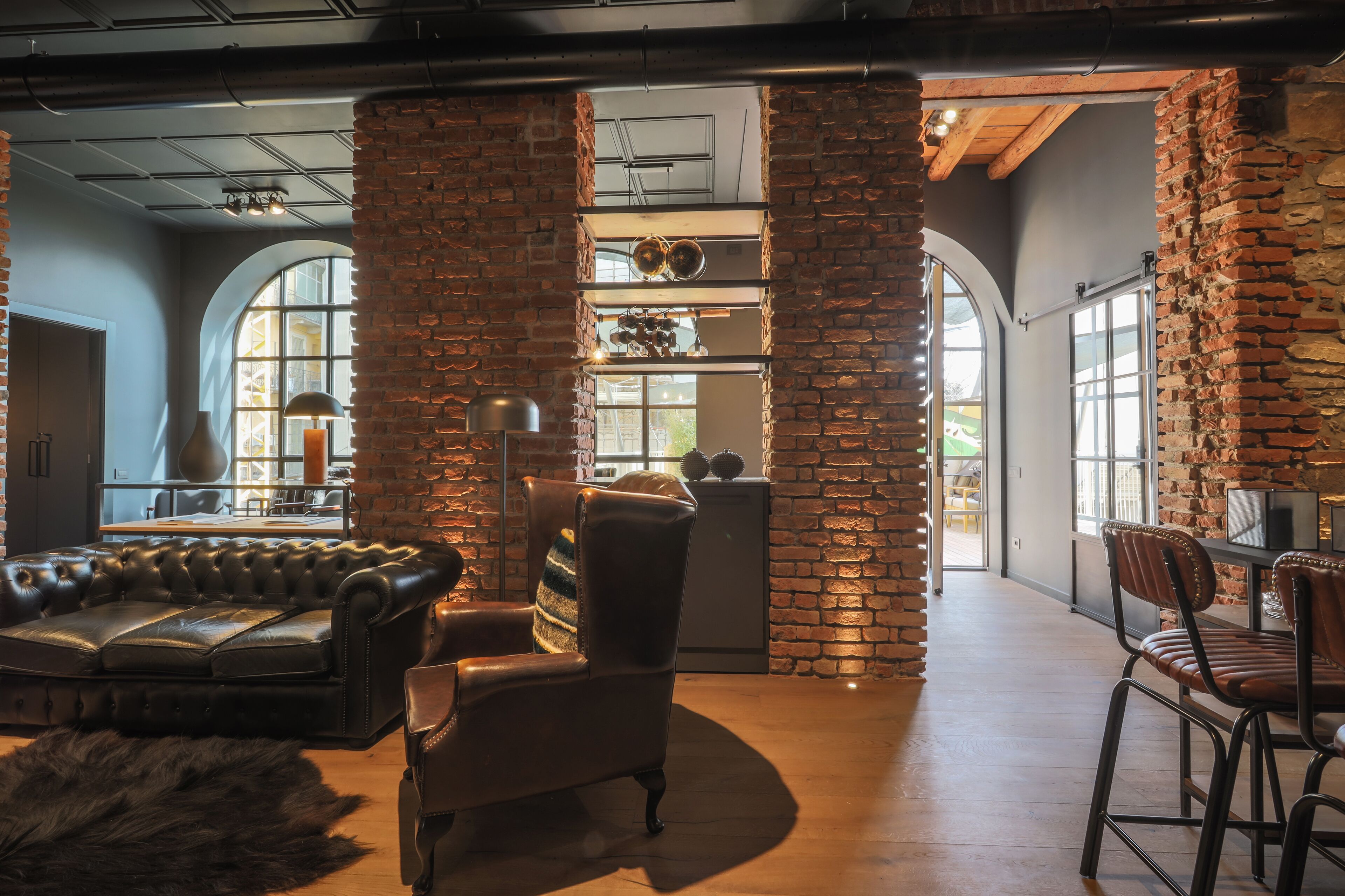 A spacious room with a leather sofa, a wooden chair, and a fur rug. There are brick walls and large windows allowing natural light.