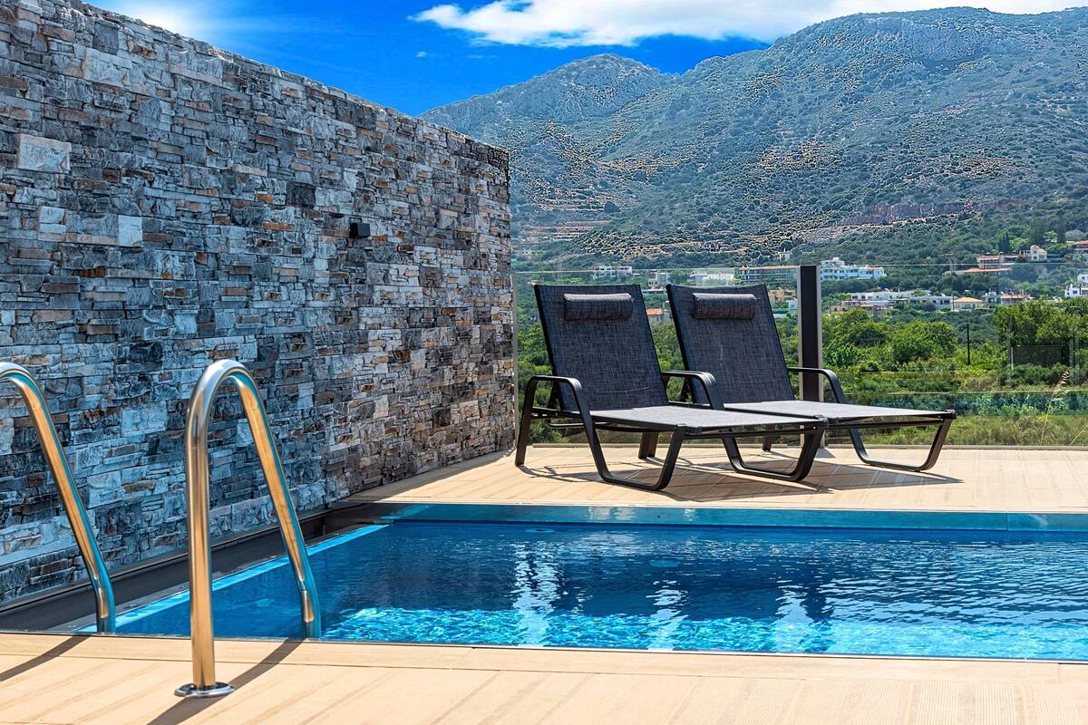 A pool area with stone walls, two lounge chairs, and a view of mountains.