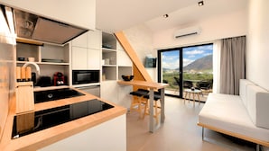 Eine moderne Küche mit einem schwarzen Induktionskochfeld, wei ßen Schränken und einem eingebauten Backofen. Es gibt einen Essbereich mit einem Holztisch und Stühlen sowie ein Sofa. Ein großes Fenster bietet Blick auf eine gebirgige Landschaft.
