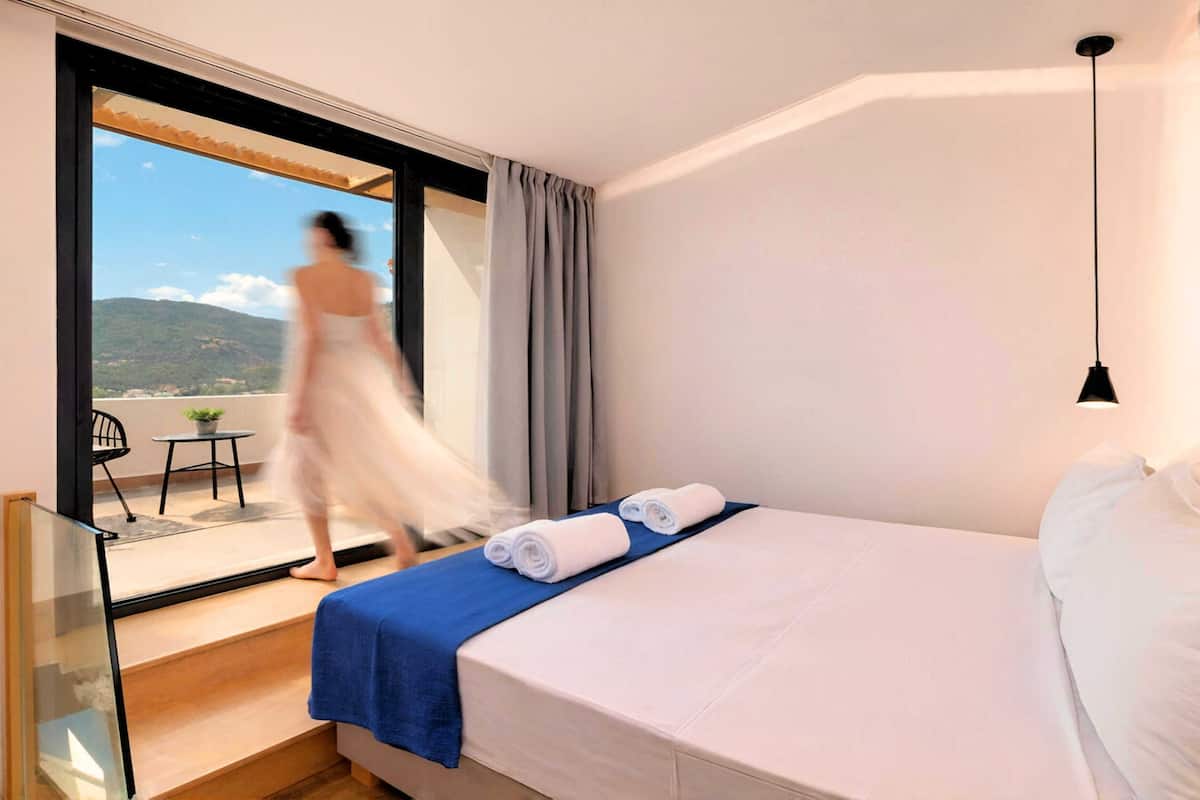 A hotel room with a bed, a balcony with a table and chair, and a view of the outdoors.