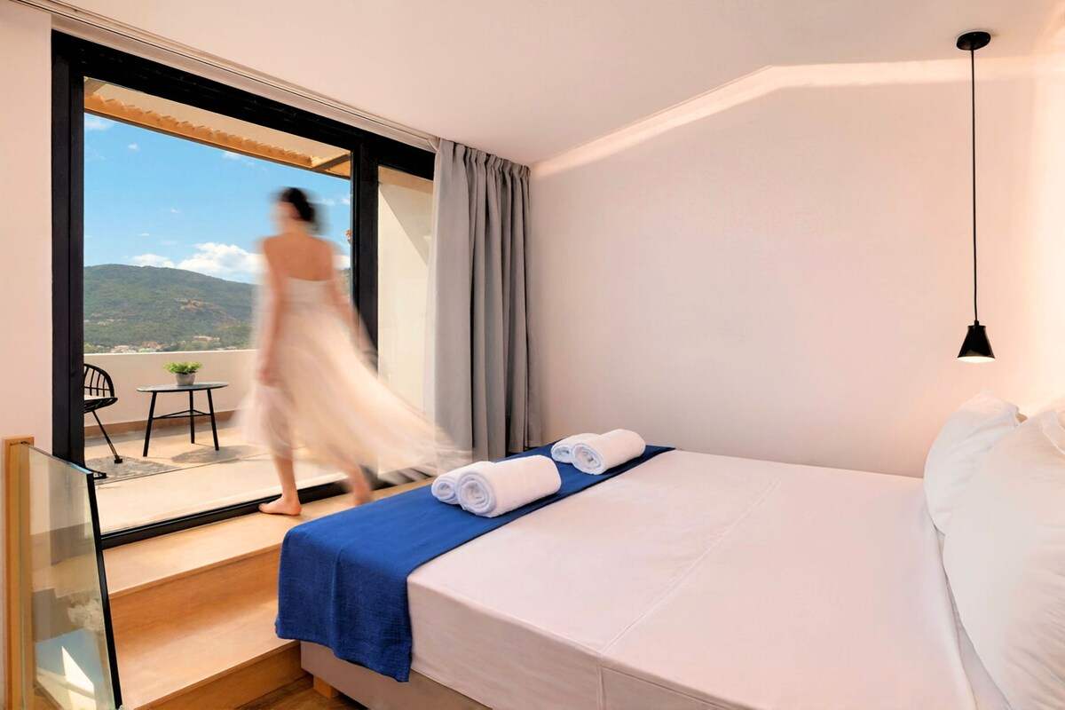 A hotel room with a bed, a balcony with a table and chair, and a view of the outdoors.