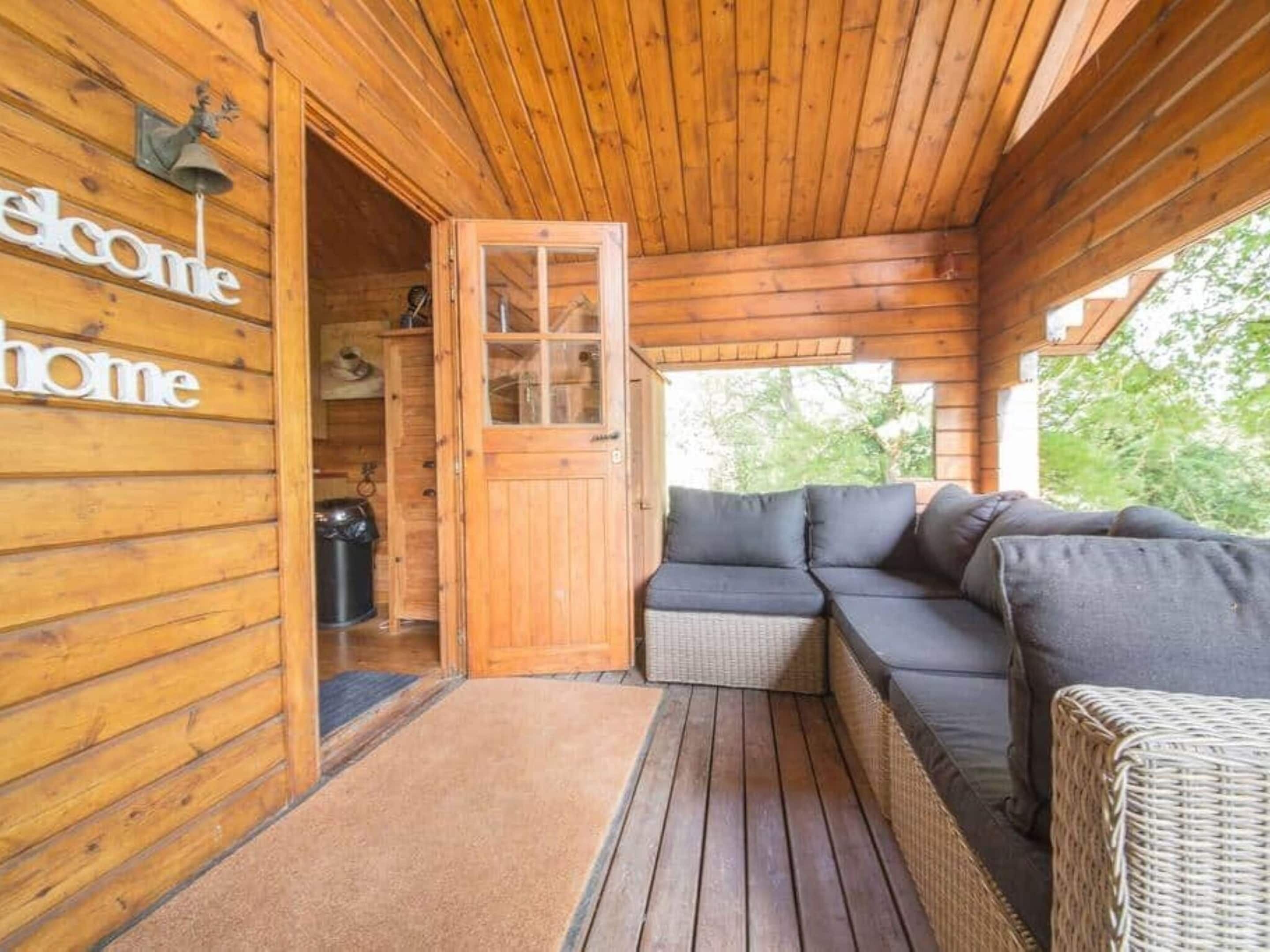 A wooden cabin with a welcoming sign, a glass door, a wicker sofa, and a wicker chair.