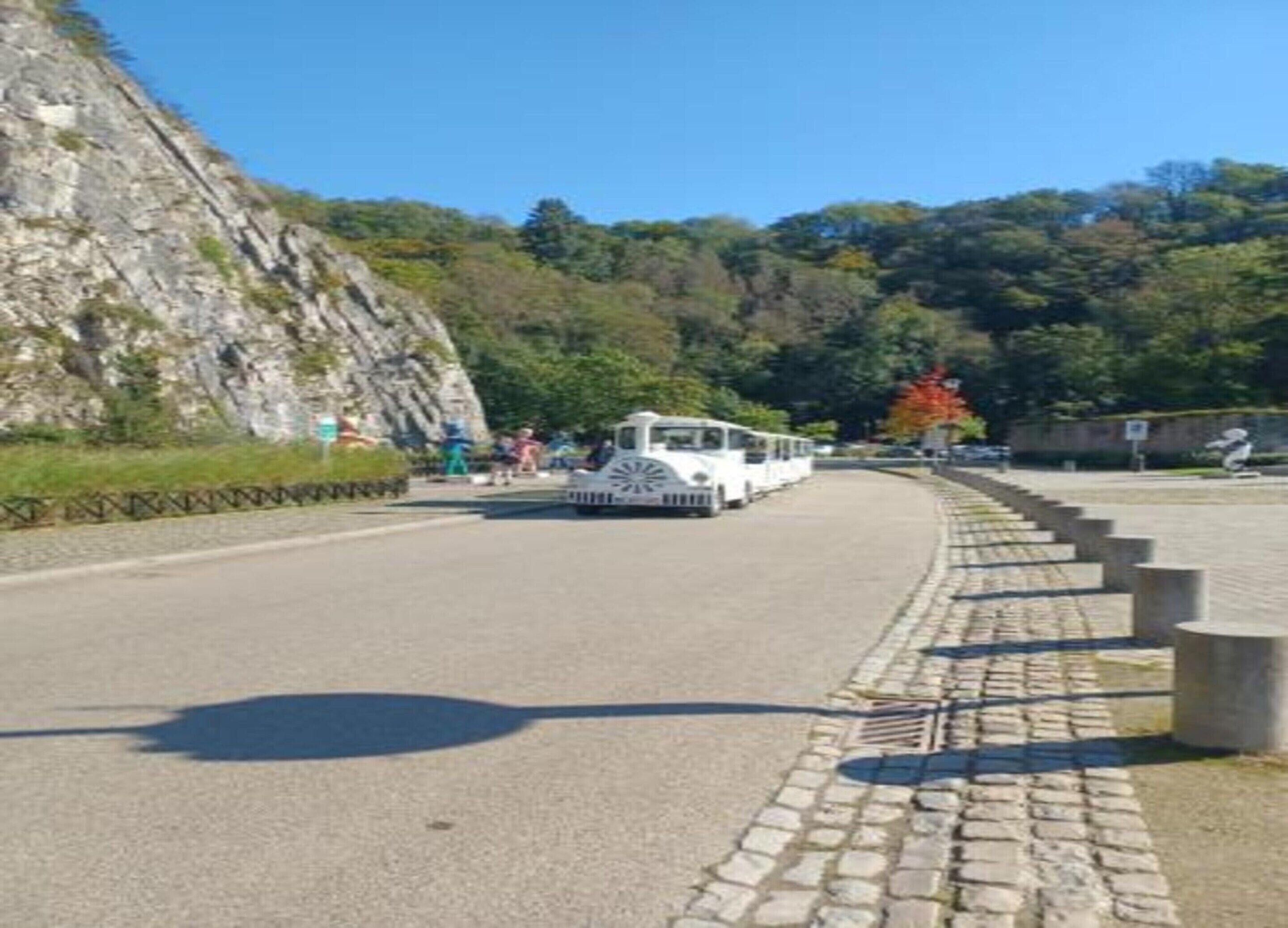 A white tram on a paved road with a stone pathway on the side, surrounded by greenery and a rocky hillside.