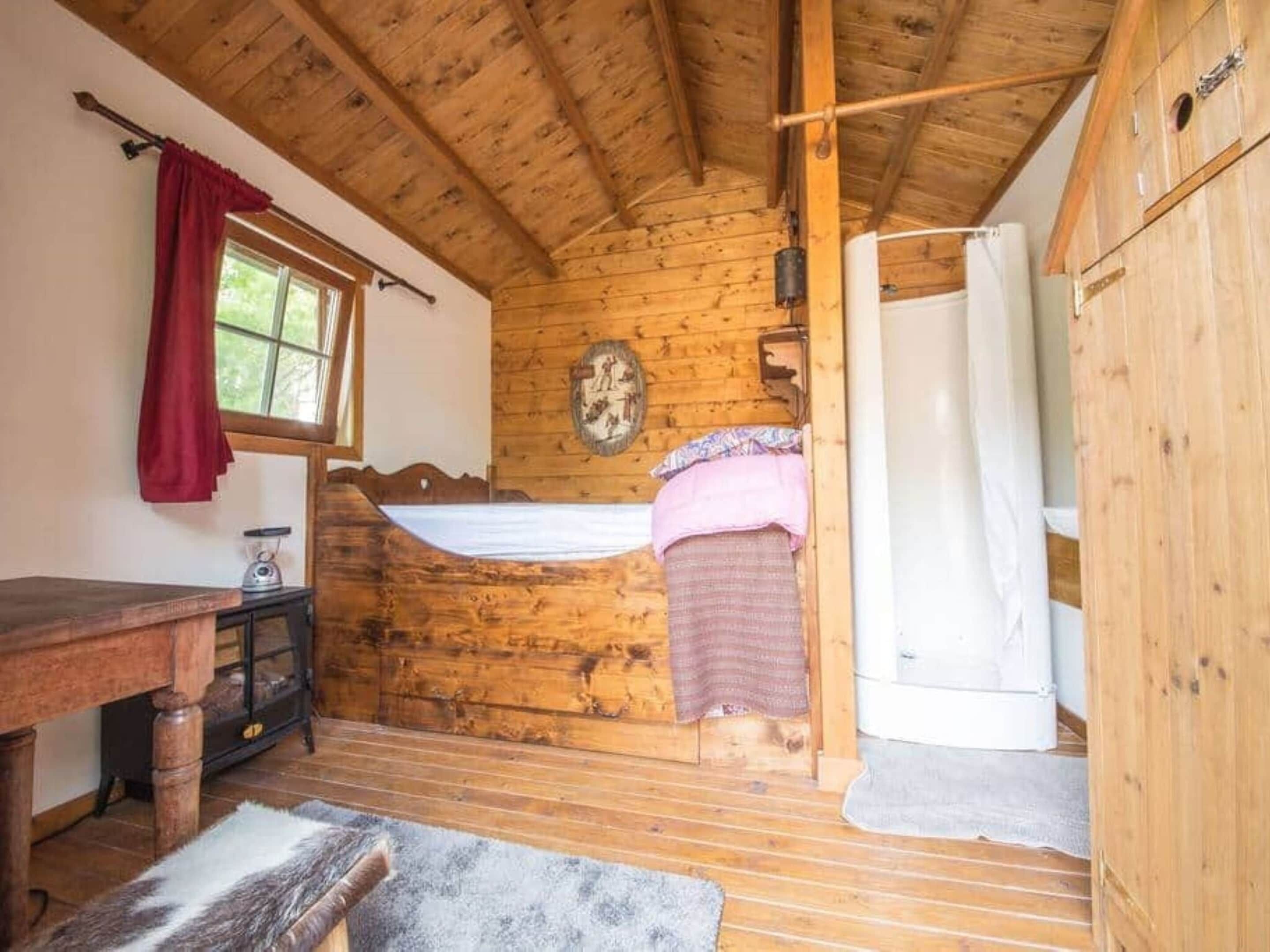 A wooden cabin with a bed, a small table, a window with a red curtain, and a white shower area.