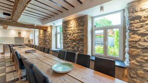Une longue table à manger en bois, entourée de chaises, un mur en pierre et une fenêtre donnant sur un paysage verdoyant.
