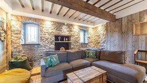 Un salon chaleureux avec un mur en pierre, des poutres en bois, un canapé d’angle gris, une table basse en bois et un fauteuil en forme de haricot vert.
