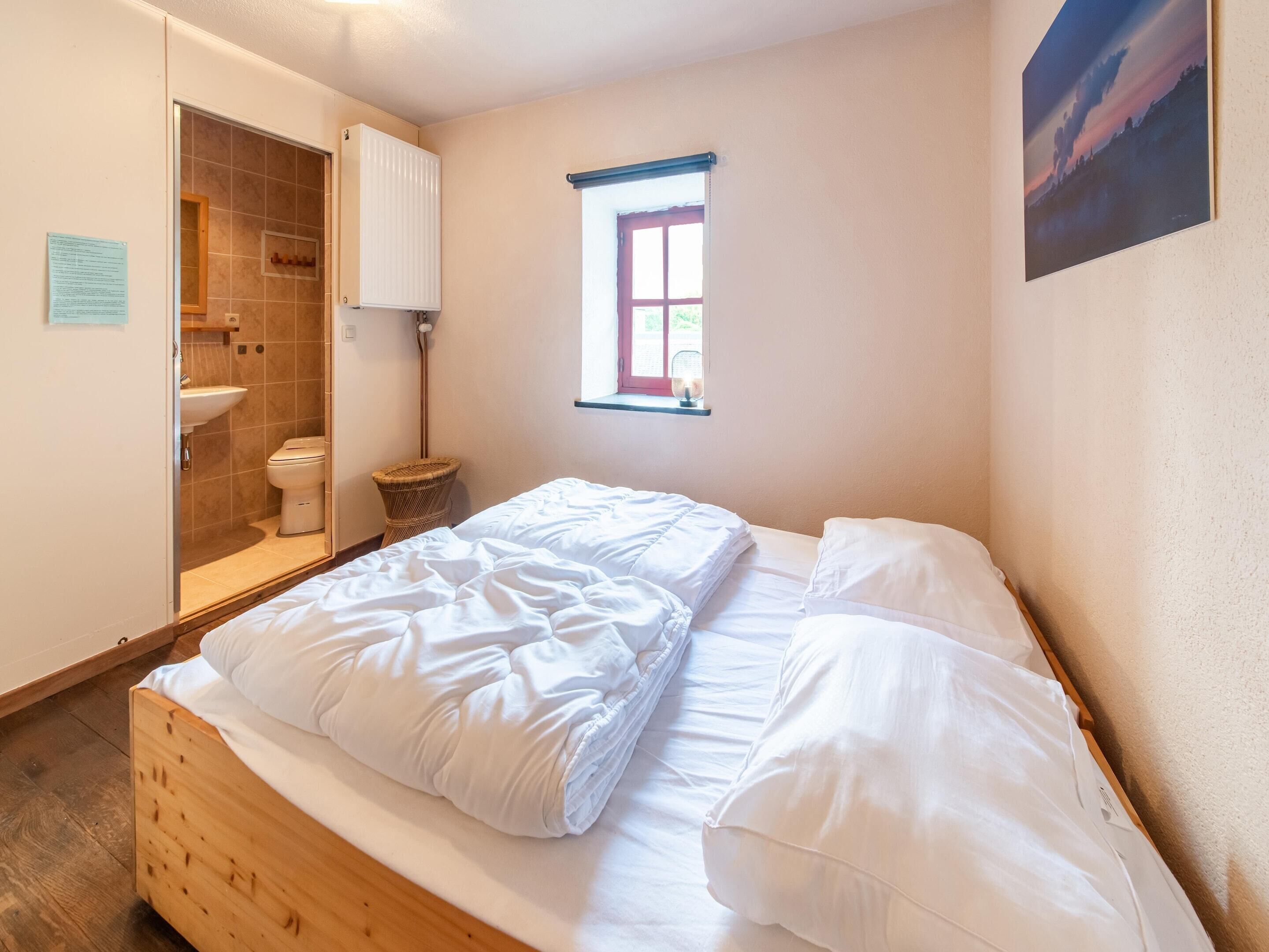 Un dormitorio con cama de madera, ventana con vistas, radiador y baño visible a través de una puerta abierta.