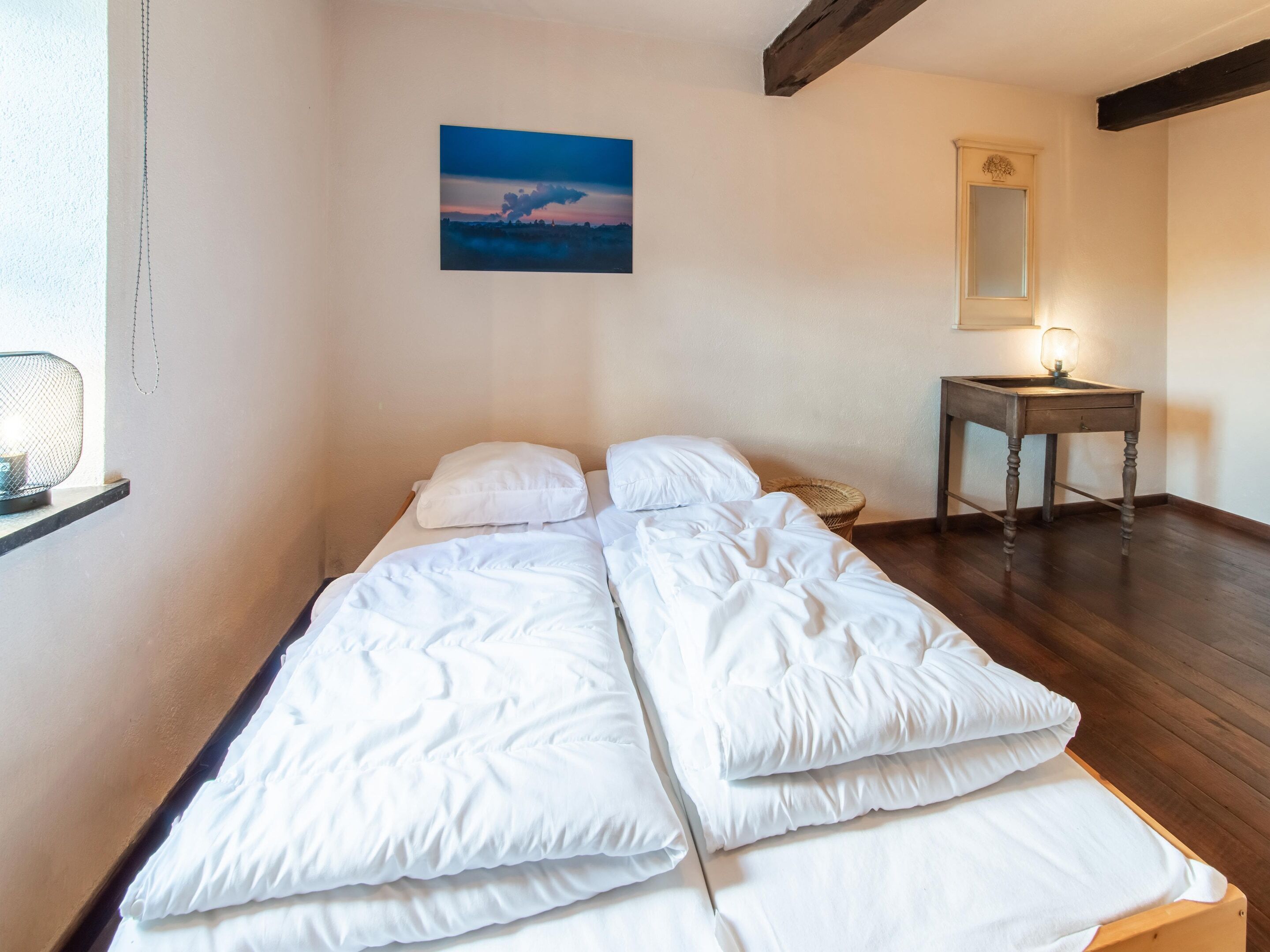 A single bed with white bedding in a room with wooden flooring, a small table with a lamp, and a painting on the wall.
