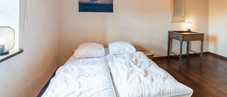 A single bed with white bedding in a room with wooden flooring, a small table with a lamp, and a painting on the wall.