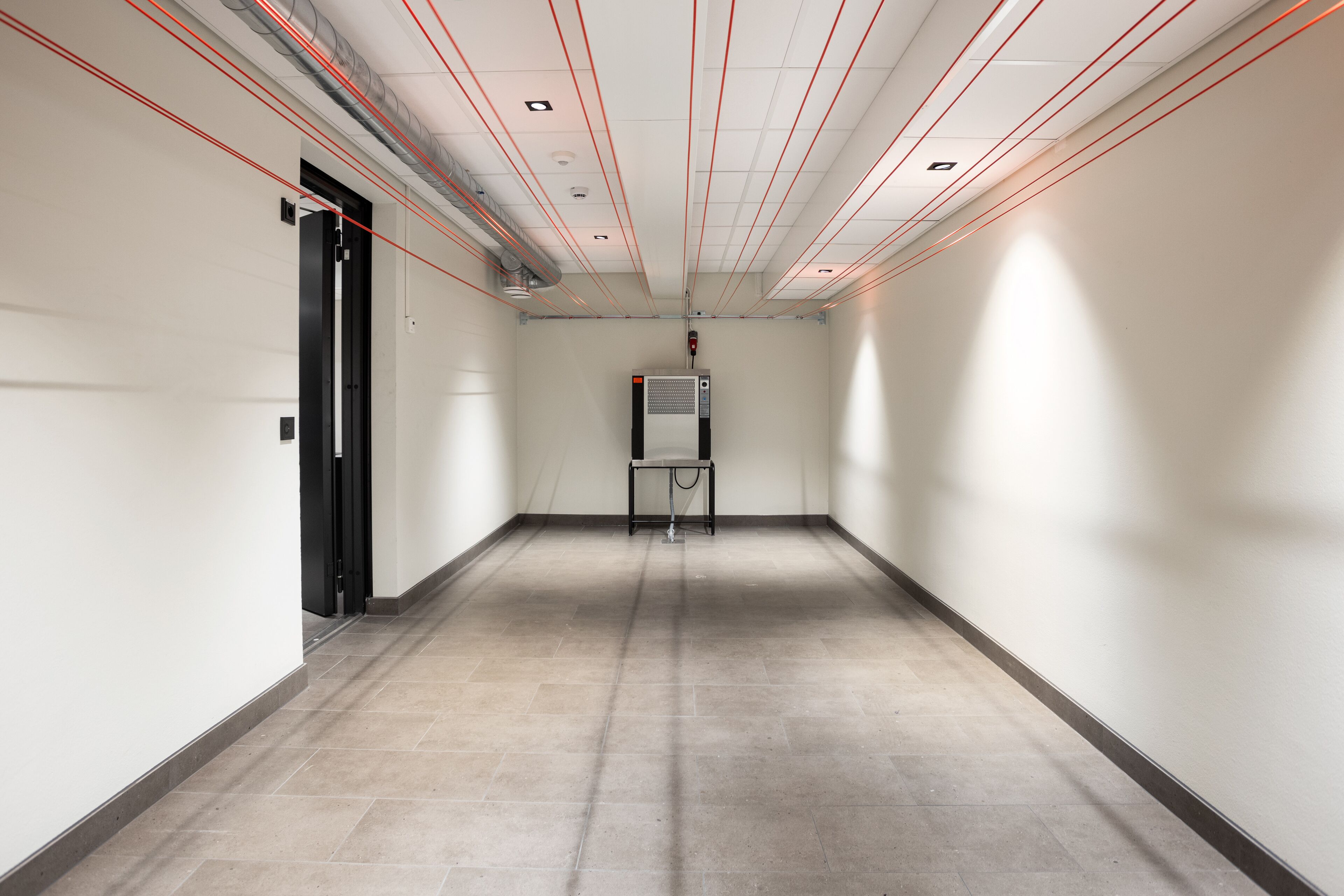 A hallway with a tiled floor, a black door on the left, and a wall-mounted air conditioning unit in the center.