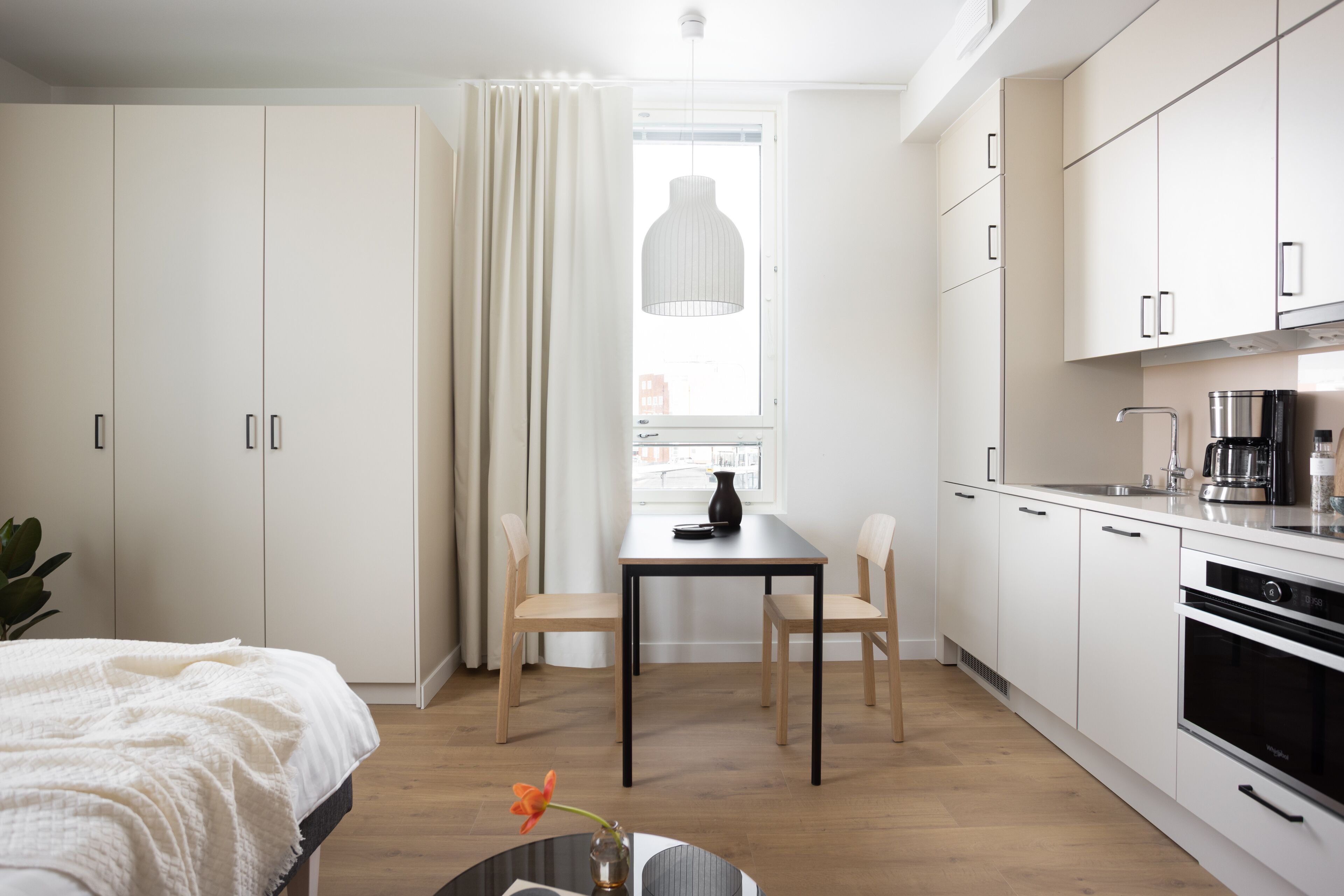 Una habitación moderna y funcional con cama, una mesa de comedor con sillas, una cocina y un ventanal con cortinas.