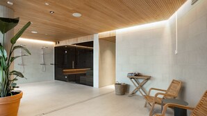 A modern spa area with wooden flooring, a large potted plant, wooden chairs, a small table, and a glass-enclosed shower area. - Noli Malmi (Helsinki)