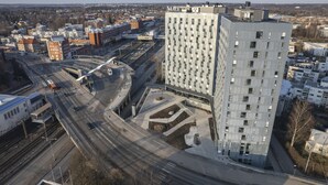 A high-rise building with a grid-like facade, situated in an urban area with multiple roads and a train track nearby. - Noli Malmi (Helsinki)
