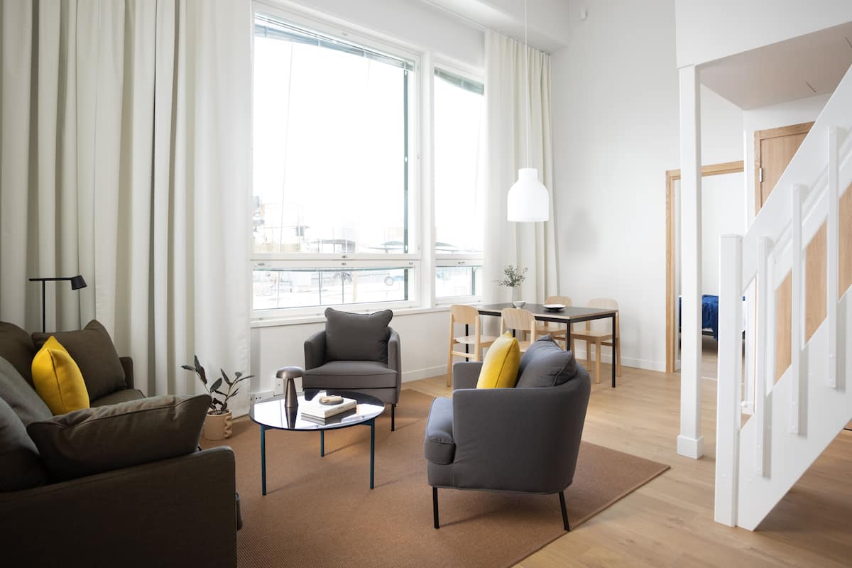 Uma sala moderna com sofá, poltrona e mesa de centro, uma área de jantar com mesa e cadeiras, e uma escadaria que dá acesso a outro andar.
