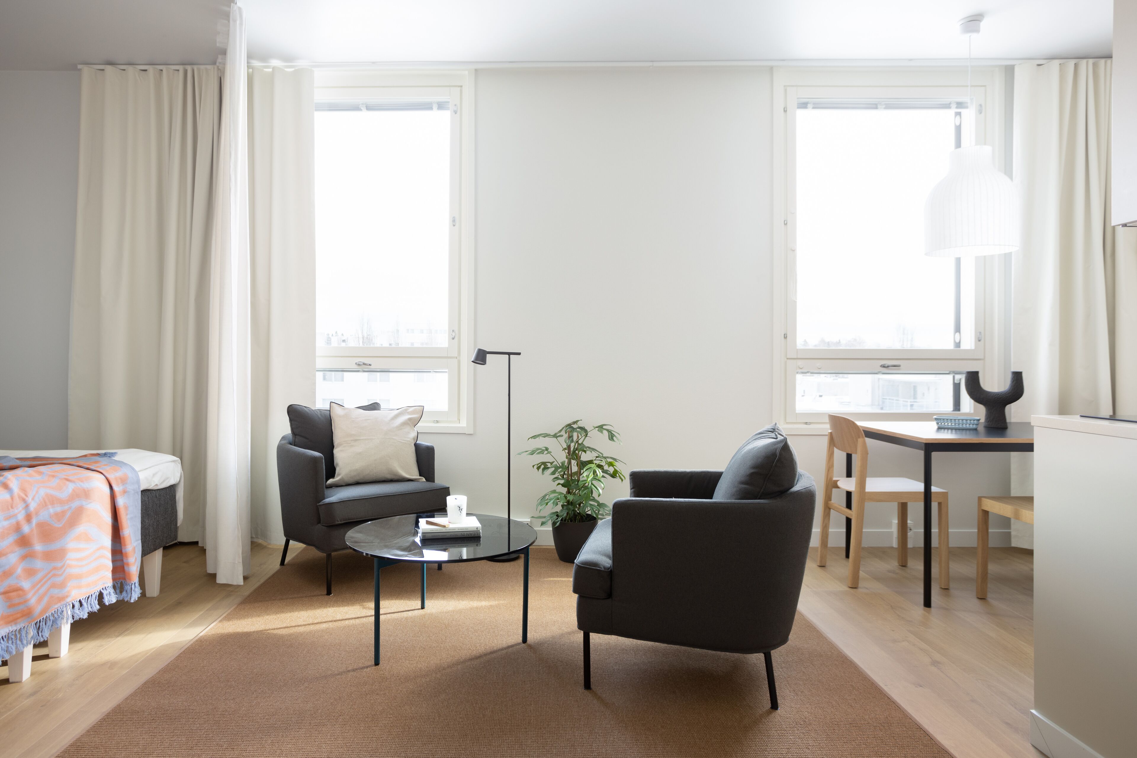 Ein modernes Wohnzimmer mit Sofa, Sesseln, einem Couchtisch und einem Essbereich mit Tisch und Stühlen. Eine Zimmerpflanze und eine Lampe sind ebenfalls vorhanden.