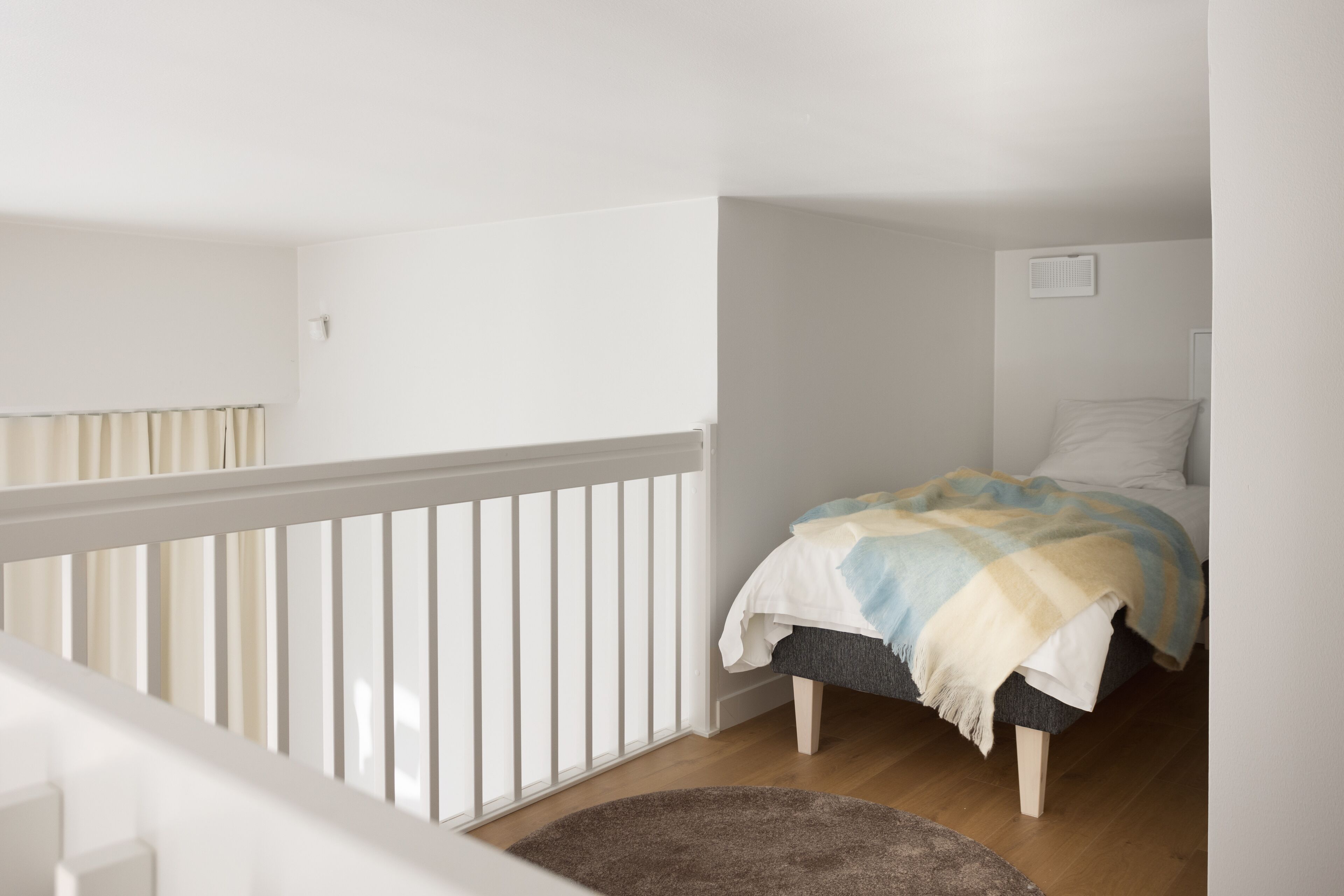 A bedroom with a bed, a window with curtains, a white railing, and a wooden floor.