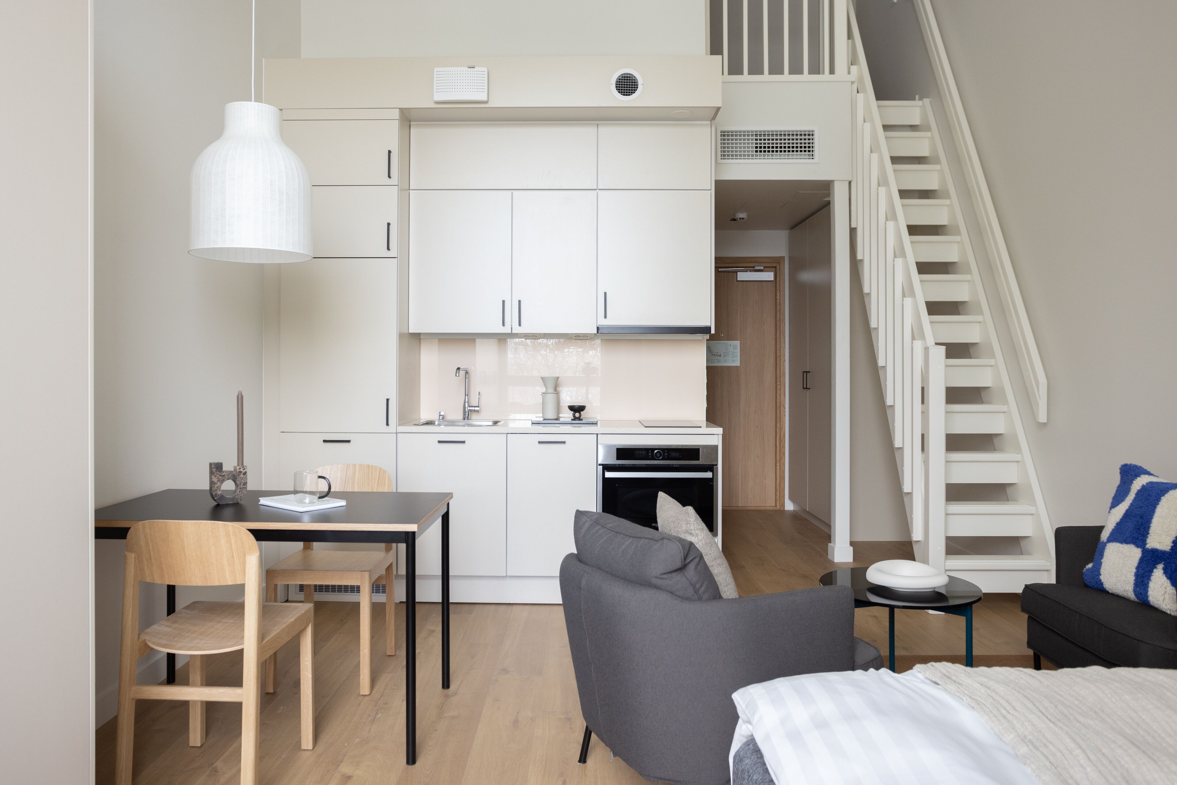 A modern kitchen with white cabinets, a dining area with a table and chairs, a sofa, and a staircase leading to an upper level.