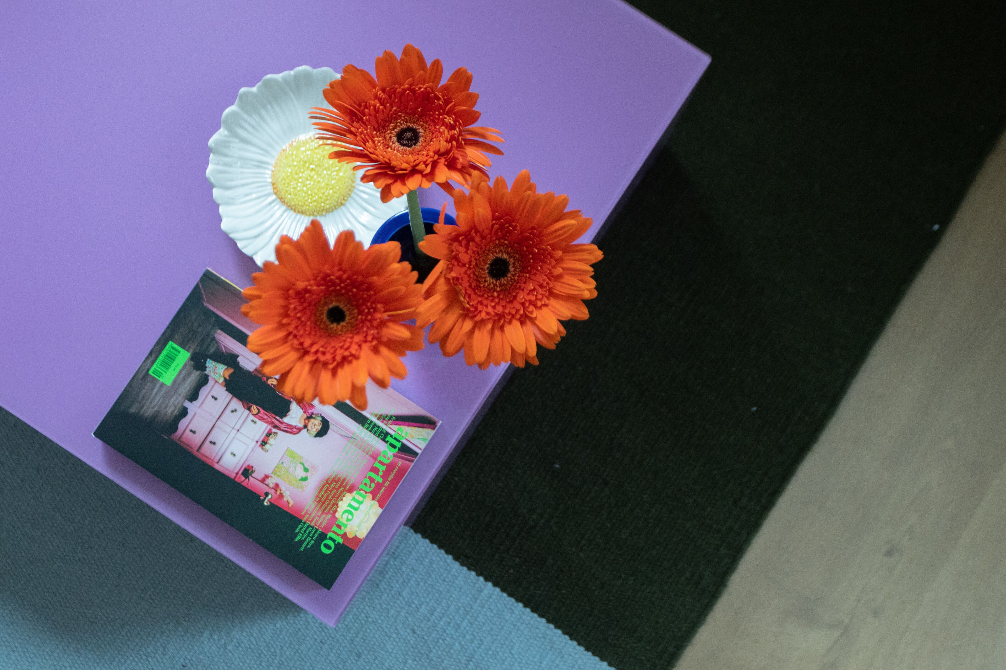 Una mesa morada con tres flores anaranjadas y una revista que muestra un interior de cocina.